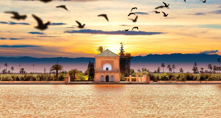 Paviljoen omringd door water met vogels die overhead vliegen bij zonsondergang.