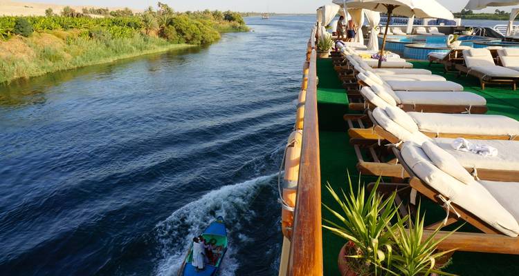 Vue depuis le pont ensoleillé d'une croisière sur le Nil bordé de chaises longues, avec un petit bateau à moteur qui file le long du fleuve en bas.