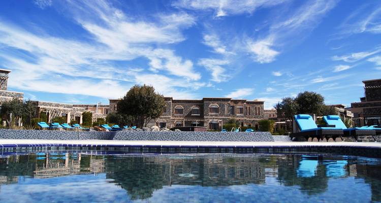 Upscale desert-style resort with turquoise loungers reflected in a still swimming pool beneath a vivid blue sky.