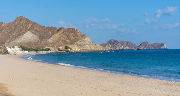 Quiet sandy beach edged by rugged brown mountains and the turquoise Arabian Sea under a clear sky in Oman.