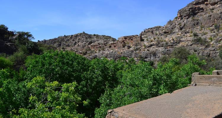 Rocky mountain slope dotted with abandoned stone houses above a lush green valley in Oman.