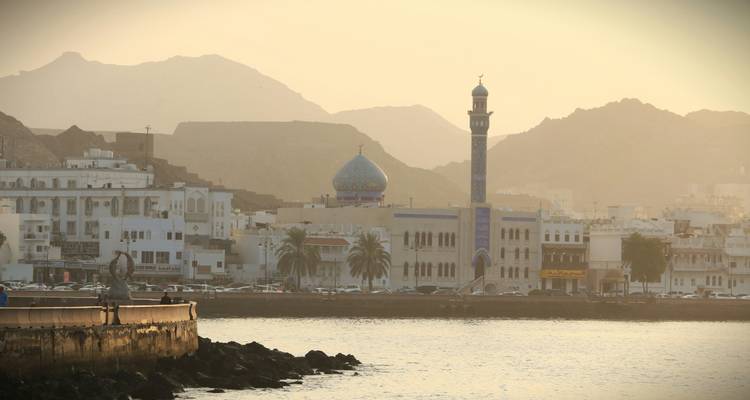 Lumière dorée et douce du matin sur le front de mer de Mascate avec un minaret de mosquée aux carreaux bleus et des montagnes escarpées en arrière-plan.