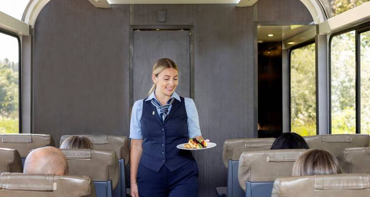 Un contrôleur de train souriant en uniforme élégant sert une collation dressée sur assiette aux passagers assis dans un wagon de luxe.