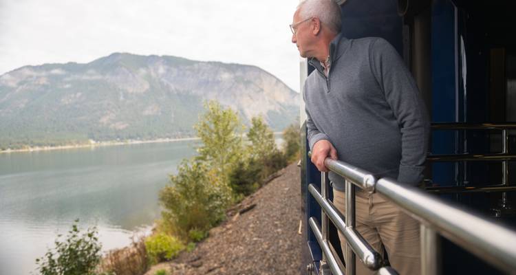 Un homme âgé profite de l'air frais sur un quai de train extérieur, contemplant un lac calme et des montagnes boisées.