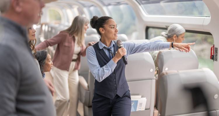 Un hôte de train en gilet et cravate pointe par la fenêtre avec un microphone tandis que les passagers se rassemblent pour regarder la vue.