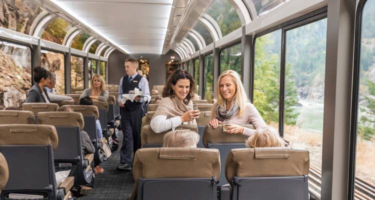 Spacieux wagon de train où les passagers discutent et sirotent des boissons pendant que les parois du canyon et la rivière défilent à travers les fenêtres.