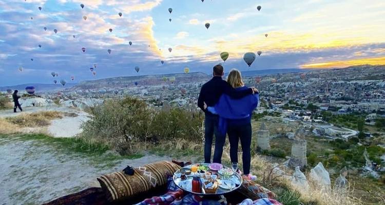 Paar beobachtet Heißluftballons bei Sonnenaufgang mit einem Picknick-Setup auf einer Klippe.