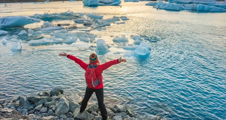 Personne debout sur des rochers avec les bras étendus, contemplant un lac glacé avec des icebergs flottants.