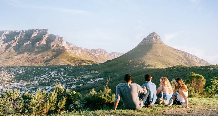 Grupo de amigos sentados en una ladera con vista a Ciudad del Cabo, Table Mountain y Lion's Head al atardecer.