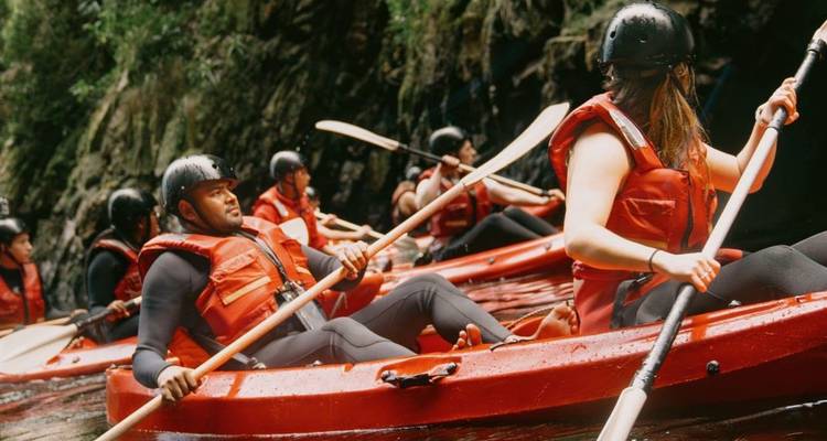 Group in red kayaks paddling through a narrow forested gorge wearing helmets and vests.