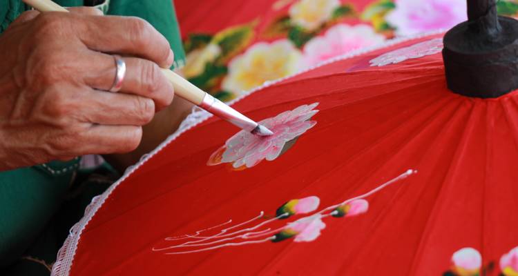 Close-up van een hand die een levendig ontwerp schildert op een rode paraplu, waarbij traditioneel vakmanschap wordt getoond.