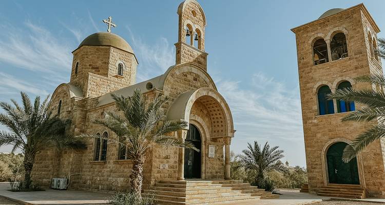 Iglesia de piedra con campanario y cúpulas rodeada de palmeras bajo un cielo azul brillante de Jordania.