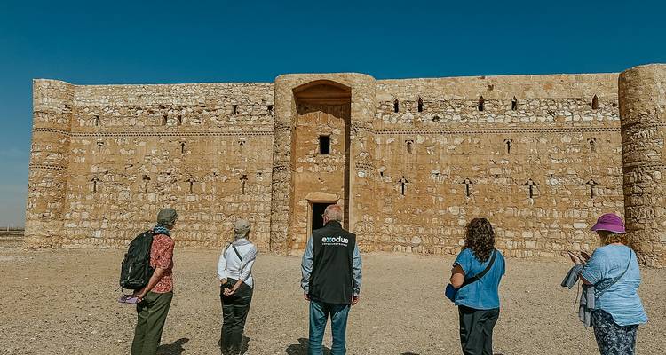 Un pequeño grupo de turistas camina a través de una llanura desértica hacia un enorme castillo del desierto omeya de paredes planas.