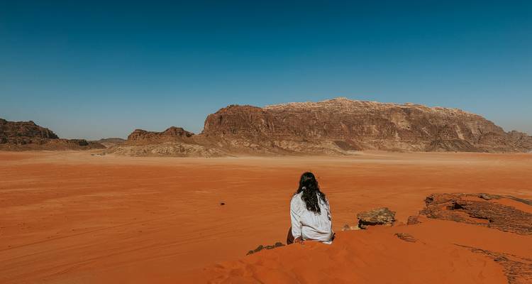 Un viajero solitario se sienta en la cima de una duna de arena roja contemplando el vasto desierto y los acantilados de arenisca de Wadi Rum.
