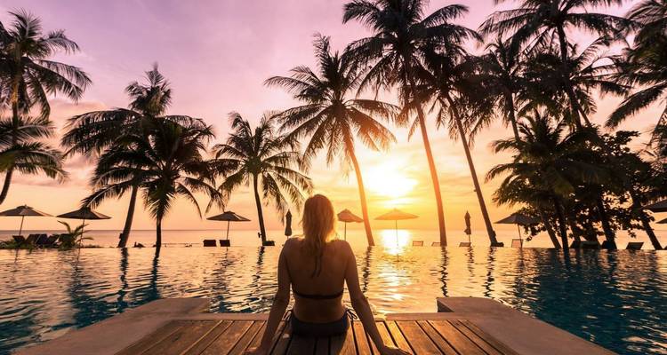 Woman sitting on a deck overlooking a pool with palm trees at sunset.