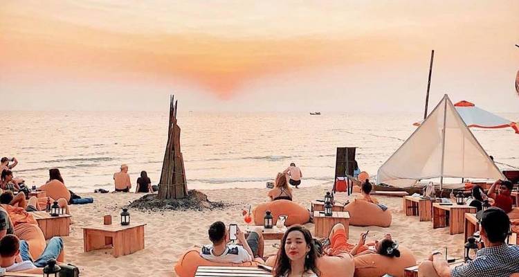 Crowds relax on beanbags and low tables at a lively beach bar during a vivid orange sunset.