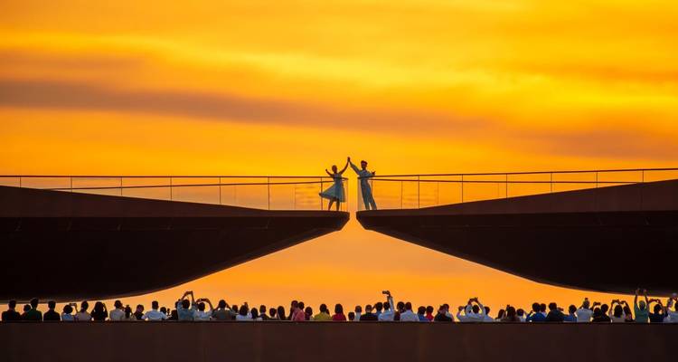 Silhouetted visitors line an elegant modern bridge between two large structures against a glowing golden sky.