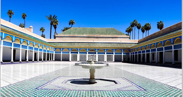 Un patio de un gran palacio con una fuente central, rodeado de columnas intrincadas y palmeras.