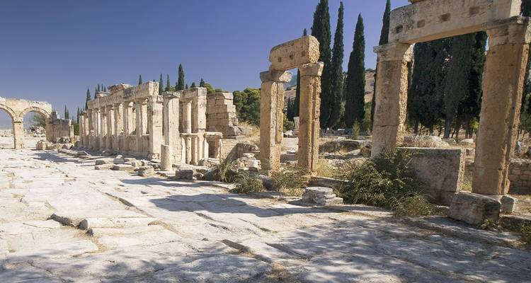 Stone colonnades and ruins of ancient Hierapolis framed by tall cypress trees on a sunny day.