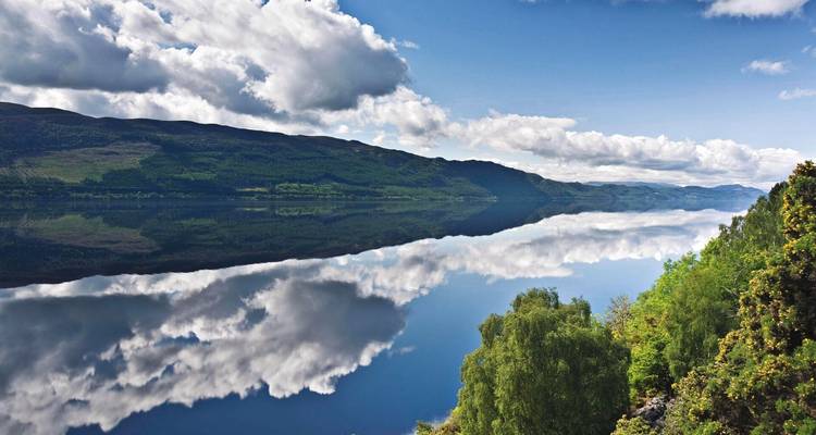 Le loch Ness, semblable à du verre, reflète les nuages et les collines vertes sous un soleil éclatant.