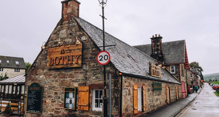 Un bâtiment en pierre rustique avec l'enseigne 'The Bothy' longe une rue de village humide.
