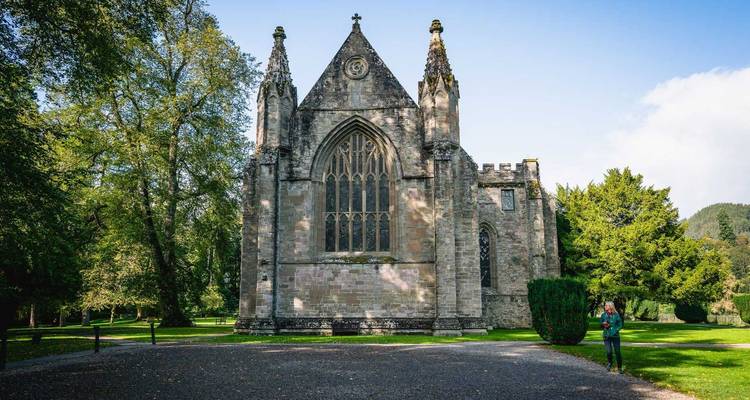 Un visiteur photographie une église gothique en pierre entourée de grands arbres.