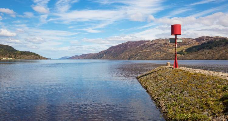 Un tronçon calme du Loch Ness avec une balise de navigation rouge et des collines vallonnées au-delà.