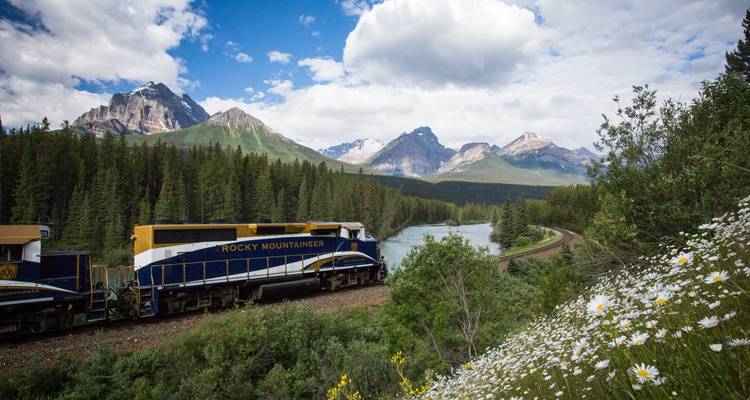 Train traveling through a lush forested area with mountains and river in the background.