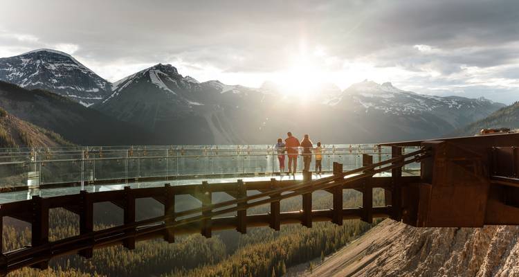 Three people standing on a glass walkway with mountains in the background at sunset.