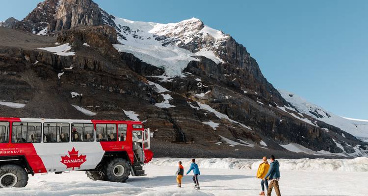 A snowcoach with people walking on a glacier with mountain backdrop.