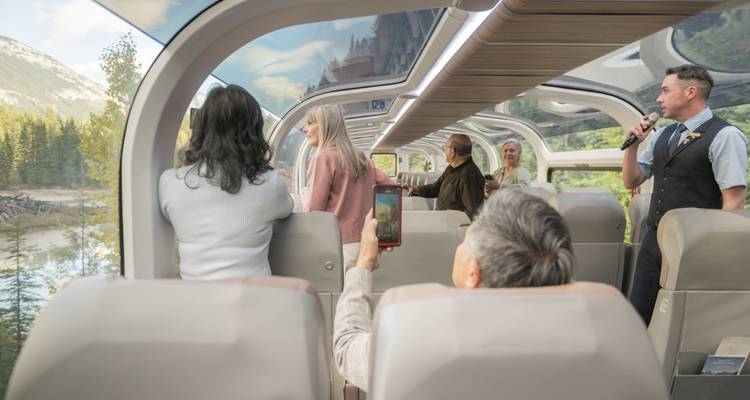 Les invités regardent vers le haut à travers le toit de verre incurvé du train tandis qu'un hôte commente le paysage.