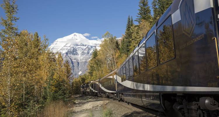 Un tren con cúpula de cristal se desliza bajo un pico nevado de montaña enmarcado por árboles otoñales.