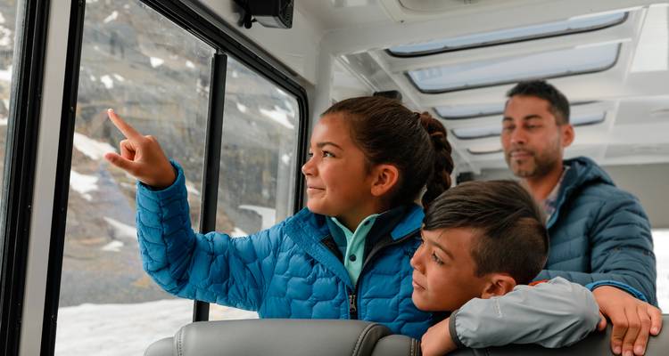 Dentro de un vehículo explorador de glaciares, los niños señalan emocionadamente el paisaje helado a través de grandes ventanas mientras un adulto los observa detrás de ellos.