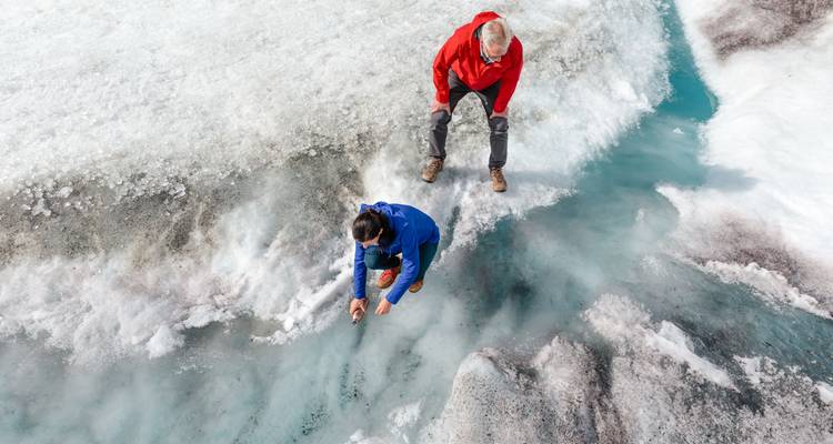 Dos viajeros con chaquetas brillantes se arrodillan sobre un arroyo de agua de deshielo azul vívido en un glaciar nevado, explorando grietas en el hielo.