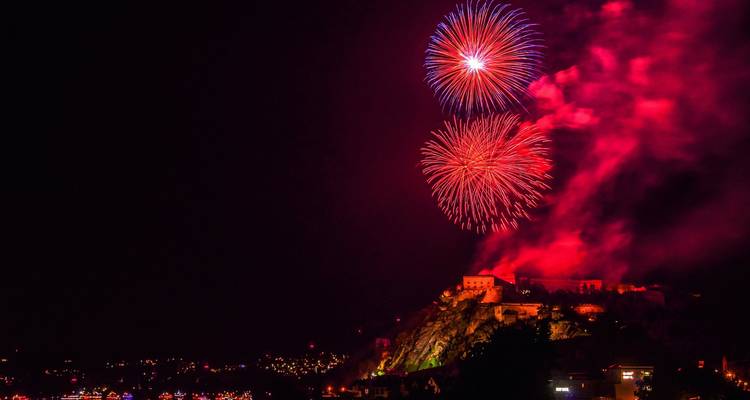 Brillantes fuegos artificiales explotan sobre una fortaleza iluminada en la cima de una colina reflejada en la noche del valle del Rin.
