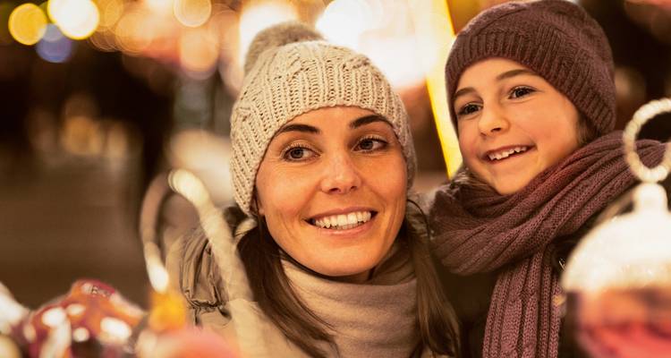 Madre sonriente y niño abrigados con gorros y bufandas tejidos disfrutan de las luces navideñas en un mercado al aire libre.