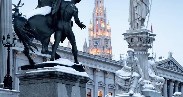 Statues couvertes de neige devant le Parlement de Vienne avec la flèche illuminée de l'Hôtel de ville dans le crépuscule hivernal