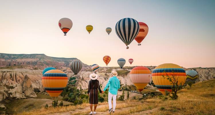 Paar hält Händchen und beobachtet Heißluftballons über felsiger Landschaft.