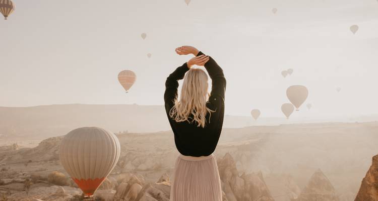 Person, die zwischen Heißluftballons steht, die über eine neblige Landschaft fliegen.