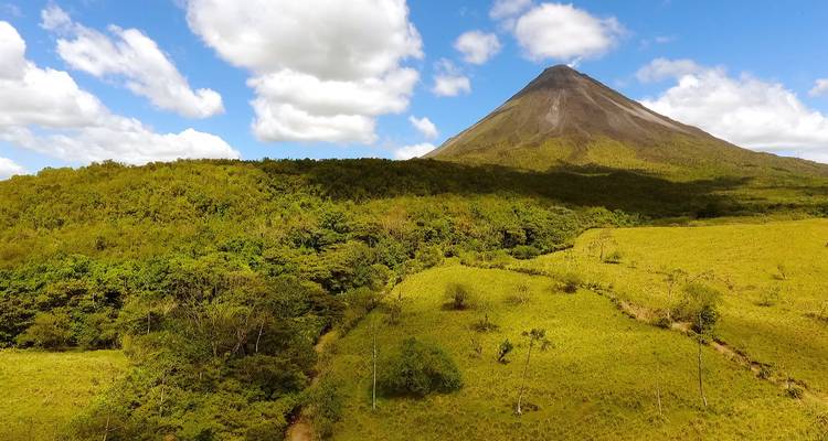 Icónico Volcán Arenal con exuberante bosque verde y cielo azul salpicado de nubes.