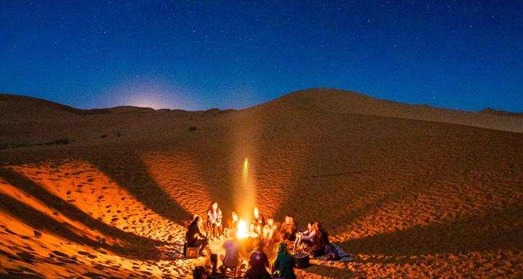 Reizigers die rond een gloeiend kampvuur zitten in de zandduinen onder een sterrenhemel.