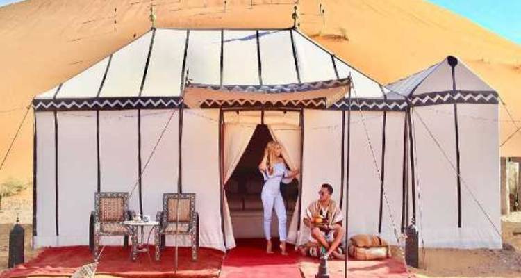 Stijlvolle canvas woestijntent tegen hoge duinen met een stel dat buiten ontspant op gevormde tapijten.