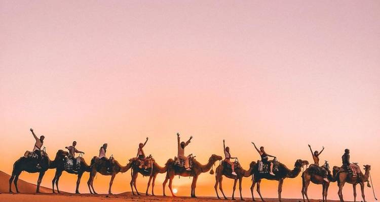 Lange rij toeristen die op kamelen rijden, afgetekend tegen een oranje woestijnhemel bij zonsondergang.