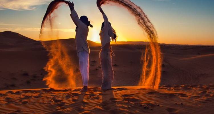 Twee vrienden gooien bogen van zand de lucht in terwijl de zon ondergaat achter golvende woestijnduinen.