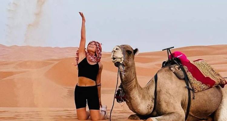 Reiziger knielend naast een zittende kameel in de Sahara, arm opgeheven naar de hemel.