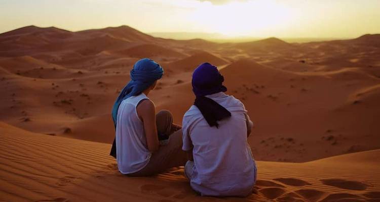 Twee reizigers zitten op een hoge duin met het gezicht naar een gloeiende zonsondergang over eindeloze zandheuvels.