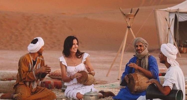 Reizigers en lokale muzikanten gezeten op tapijten in de woestijn die traditionele handtrommels bespelen bij zonsondergang.