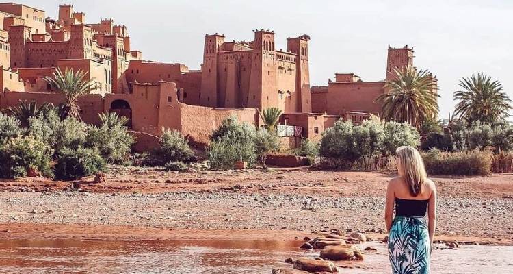 Reiziger die bij een ondiepe rivier staat en de adobe torens van Ait Ben Haddou aan de overkant bewondert.