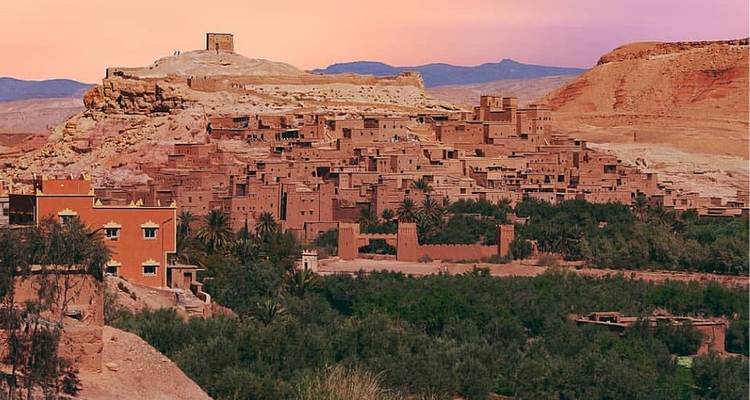 Breed uitzicht van een versterkt kasbah-dorp op een heuvel dat roze gloeit onder een pastelkleurige zonsonderganglucht.