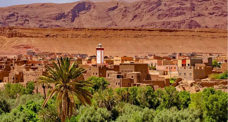 Oasestad met palmbomen en aarden gebouwen gelegen onder dorre rode bergen in felle zon.